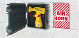 A yellow handheld air horn displayed inside an open black hard case, with a red-and-white 'AIR HORN' sign shown beside it against a light textured wall.